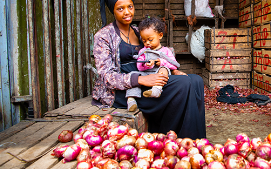 a woman holding a baby while selling onions in a market